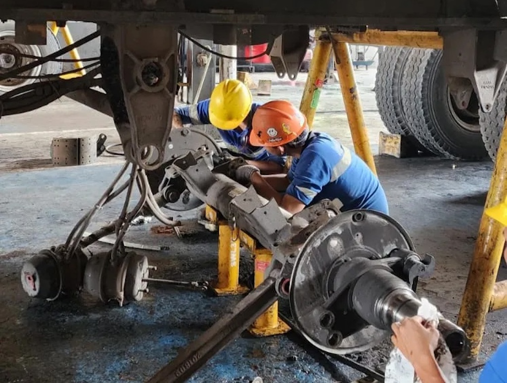A technician performs maintenance and repairs on a trailer axle system in the workshop area using complete safety equipment (Image Source: TDU)